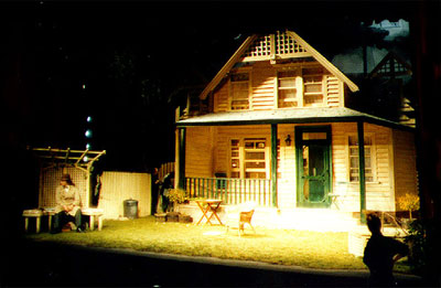 view of stage set - the exterior of an almost full-size wooden clapboard house with a porch extending from its frontage and a green-painted front door. There is a set of garden furniture in the yard in front of the porch.