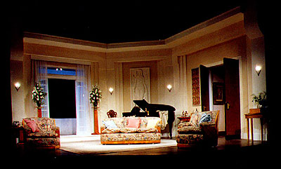 view of stage set - a classical interior with a large window and double doors. A grand piano stands upstage centre flanked by an ornate three-piece suite.