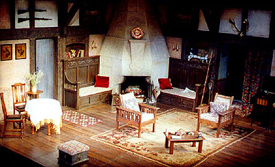 view of stage set - a large wood-panelled room with a stone fireplace upstage centre. Carved chairs and settles are grouped around the fireplace.